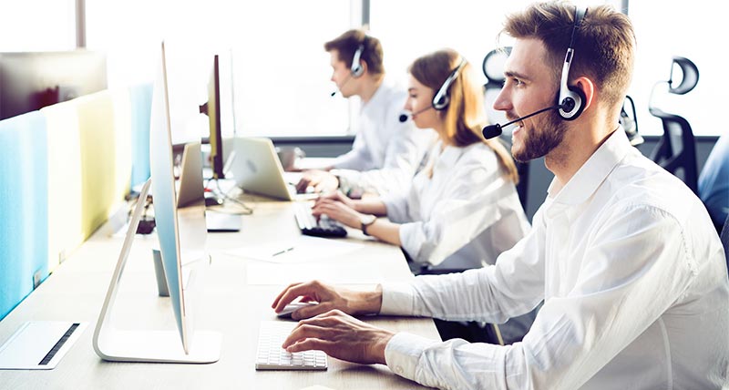 Three sales representatives sitting at a long desk typing in front of computer screens and wearing headsets